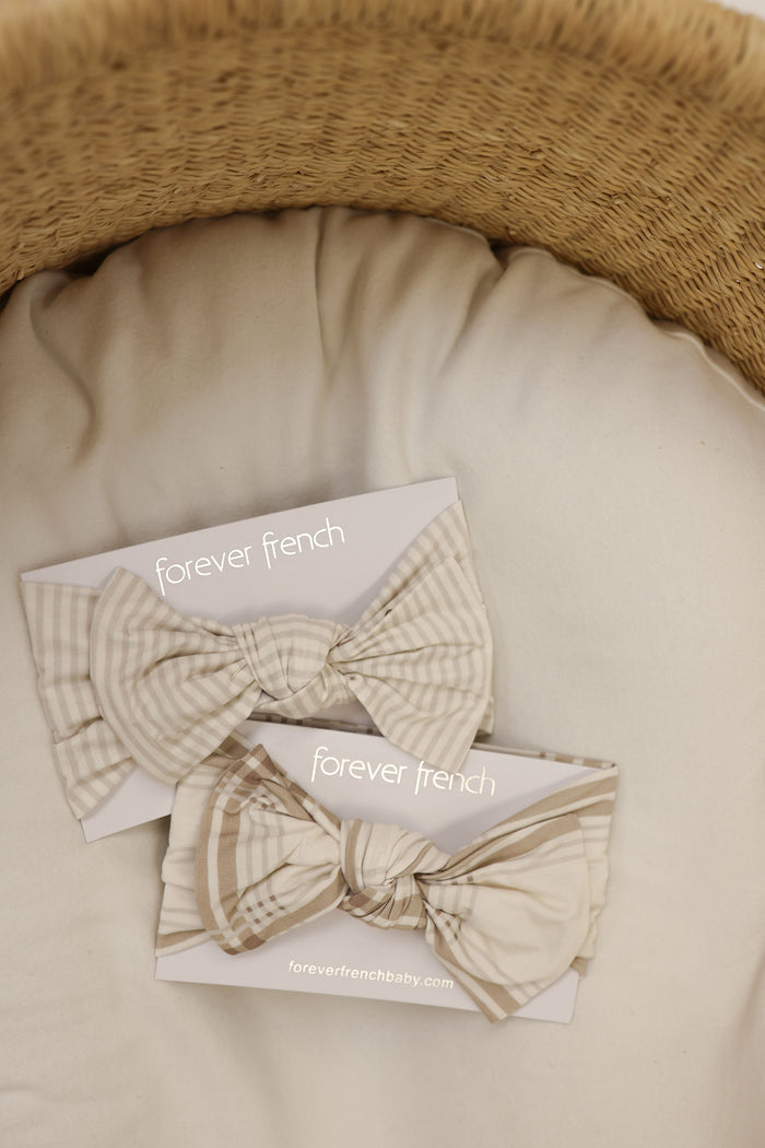 Two cream and beige Ellis Plaid fabric bows from forever french baby, designed to pair with the Bamboo Head Wrap, sit on a soft cushion in a woven basket. Each bow comes attached to a branded card.