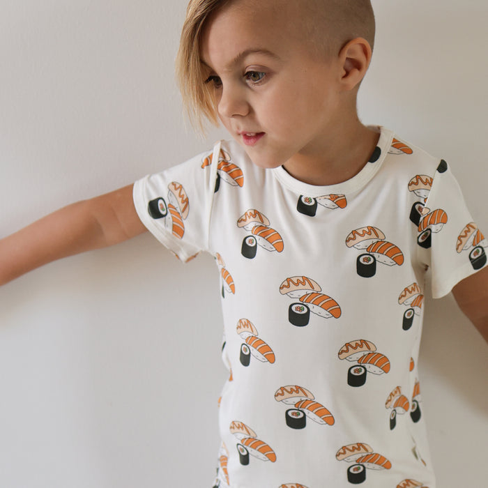A young child with short, side-shaved blonde hair wears the forever french baby Bamboo Short Set | Sushi, featuring sushi and nigiri prints on a white tee, standing against a plain white wall in soft, breathable bamboo fabric.