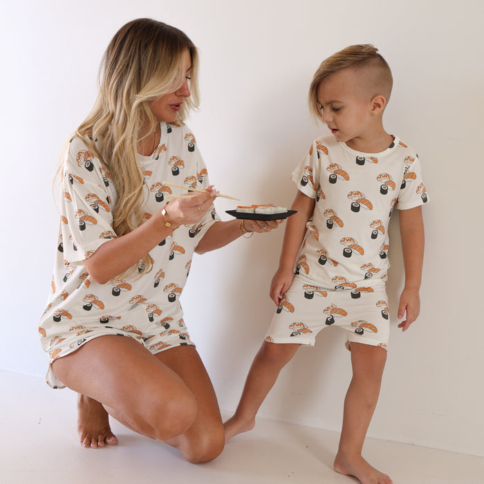 A woman and child wear matching forever french baby Bamboo Short Set | Sushi pajamas in a bright room. The woman kneels with chopsticks and sushi while the child stands nearby, both enjoying their soft, breathable bamboo outfits.