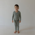 A young child with short brown hair stands barefoot on a white floor, wearing forever french baby's Bamboo Two Piece Pajamas | Lucky Micro Check—breathable, comfy sleepwear—against a plain light-colored wall.
