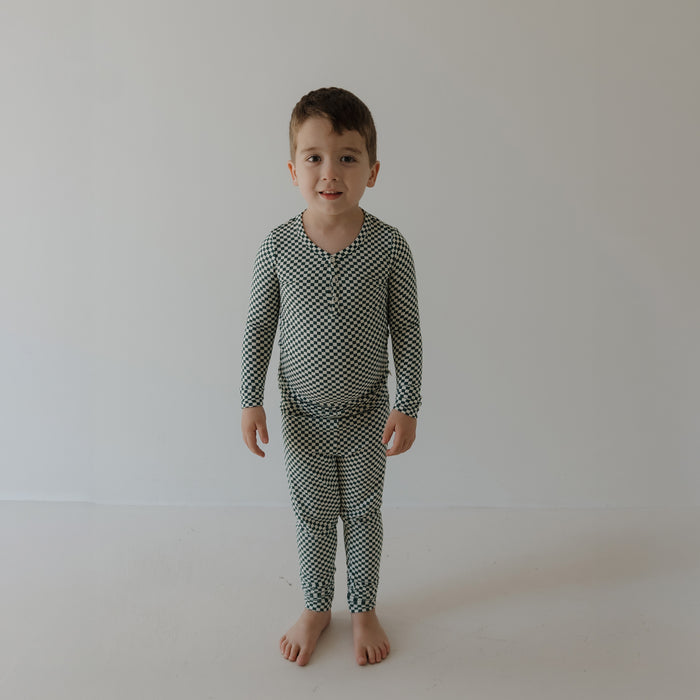 A young child with short brown hair stands barefoot on a white floor, wearing forever french baby's Bamboo Two Piece Pajamas | Lucky Micro Check—breathable, comfy sleepwear—against a plain light-colored wall.