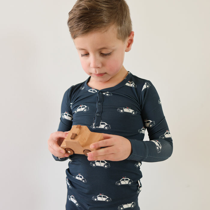 A young boy models the forever french baby Bamboo Two Piece Pajamas | Police Cruiser, featuring dark blue, car print hypo-allergenic sleepwear, while holding a wooden toy car against a plain white background.