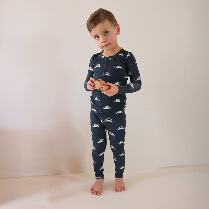 A young boy wears forever french baby Bamboo Two Piece Pajamas | Police Cruiser—navy blue with white police car patterns—as he stands barefoot against a plain light background, holding a wooden toy car. Ideal comfy, hypo-allergenic kids sleepwear.