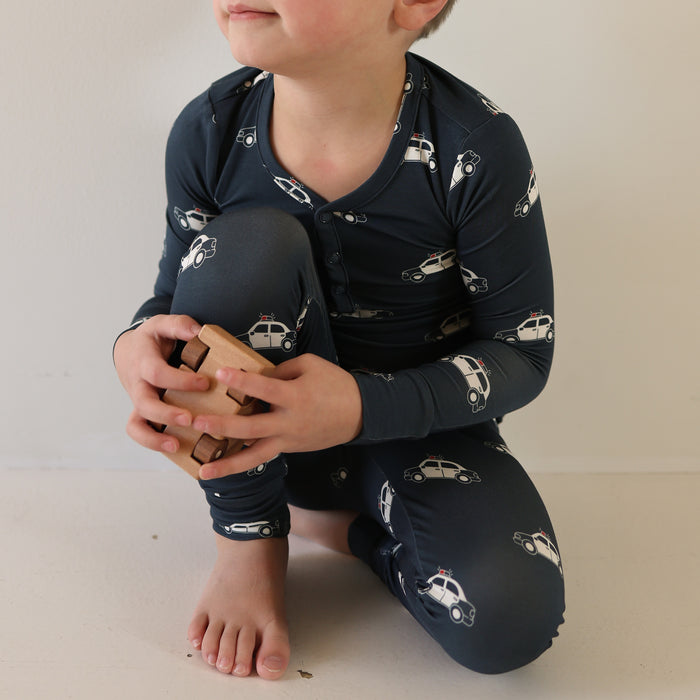 A child in forever french baby's Bamboo Two Piece Pajamas | Police Cruiser pattern crouches barefoot on the floor, holding a wooden puzzle with their face partially out of frame.