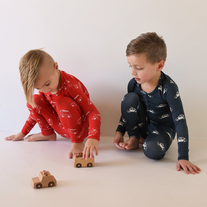 Two young children in forever french baby Bamboo Two Piece Pajamas | Police Cruiser—one in red, one in navy—sit on a white floor playing with wooden cars. Enjoy these soft, hypo-allergenic bamboo pajamas for kids.