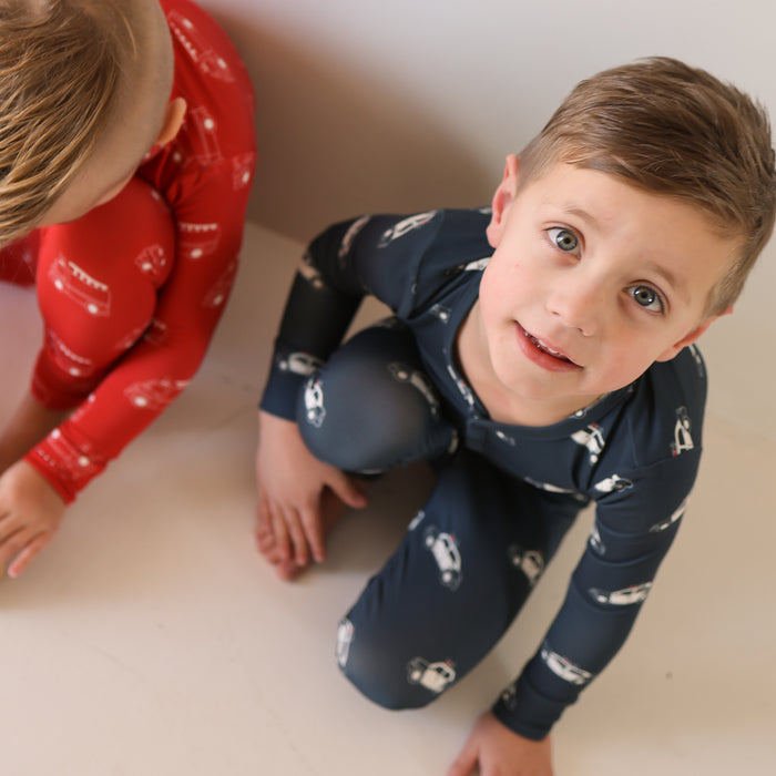 Two young children wear forever french baby Bamboo Two Piece Pajamas | Police Cruiser. One smiles at the camera in dark blue kids sleepwear, while the other, in red pajamas, is partially visible and looking downward.