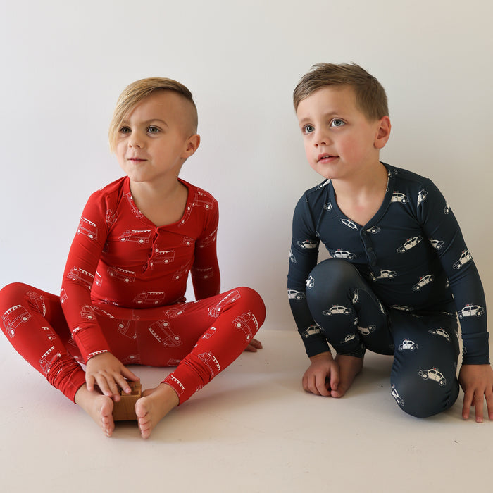 Two young children sit barefoot against a white wall in forever french baby Bamboo Two Piece Pajamas | Fire Truck. One wears red pajamas with white fire trucks, and the other dark blue pajamas. Both look slightly upward.