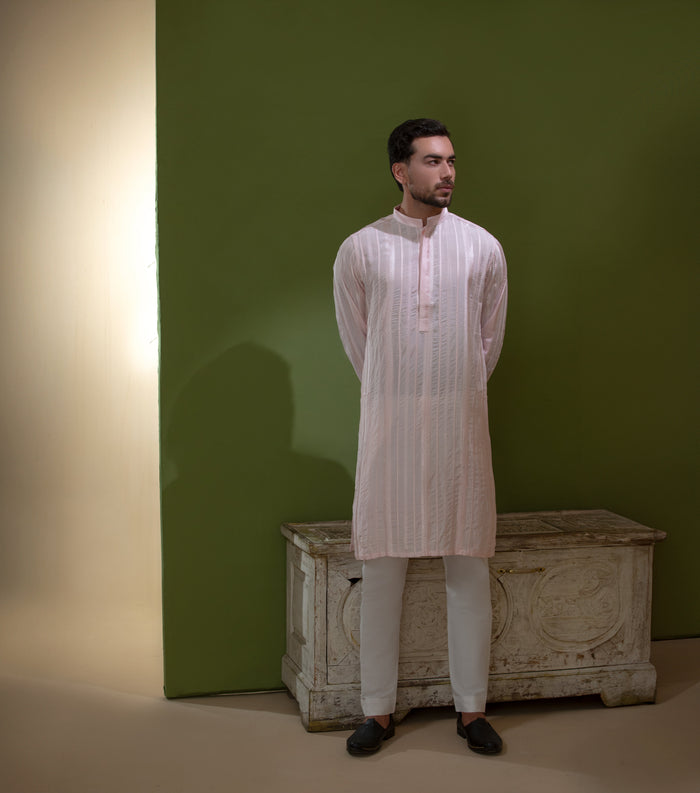Arsalan Iqbal Blush Pink Chikankari Kurta & Pyjama in Raw Silk Viscose  man wearing a light blush pink full sleeve kurta with all-over white chikankari embroidery on raw silk viscose fabric, mandarin collar with button placket, paired with matching white straight-cut pyjama, styled against an olive green wall with a rustic wooden trunk