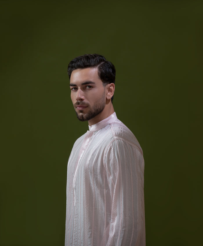 Arsalan Iqbal Blush Pink Chikankari Kurta & Pyjama in Raw Silk Viscose  man wearing a light blush pink full sleeve kurta with all-over white chikankari embroidery on raw silk viscose fabric, mandarin collar with button placket, paired with matching white straight-cut pyjama, styled against an olive green wall with a rustic wooden trunk