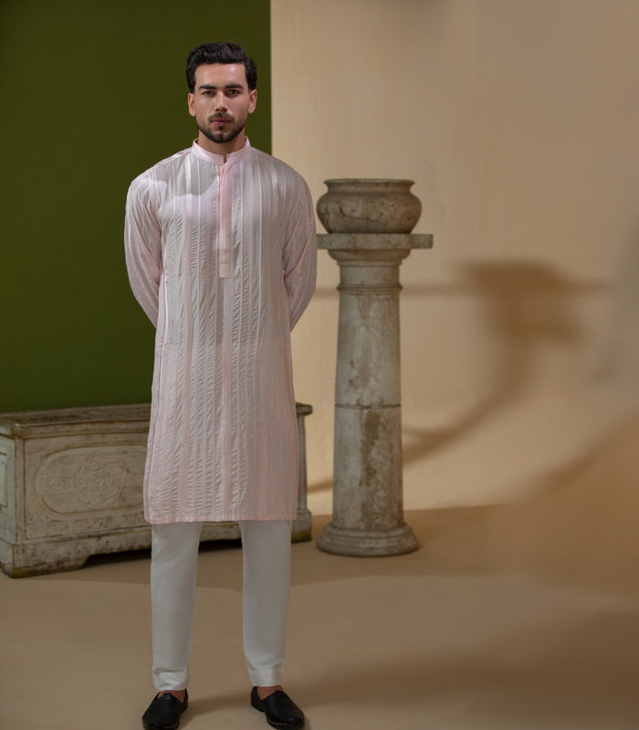 Arsalan Iqbal Blush Pink Chikankari Kurta & Pyjama in Raw Silk Viscose – man wearing a light blush pink full-sleeve kurta with all-over white chikankari embroidery on raw silk viscose fabric, mandarin collar with button placket, paired with matching white straight-cut pyjama, styled against an olive green wall with a rustic wooden trunk