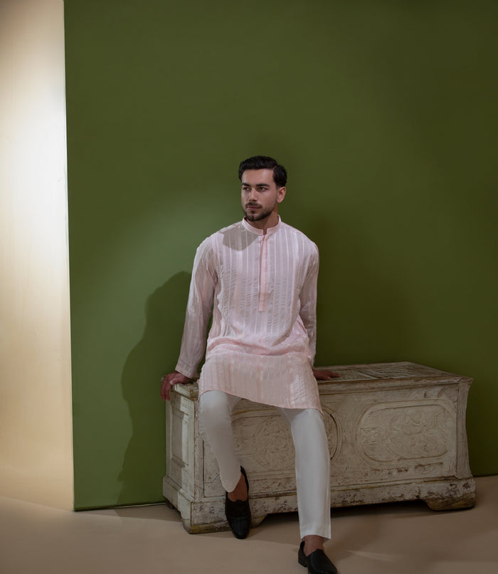 Arsalan Iqbal Blush Pink Chikankari Kurta & Pyjama in Raw Silk Viscose – man wearing a light blush pink full-sleeve kurta with all-over white chikankari embroidery on raw silk viscose fabric, mandarin collar with button placket, paired with matching white straight-cut pyjama, styled against an olive green wall with a rustic wooden trunk