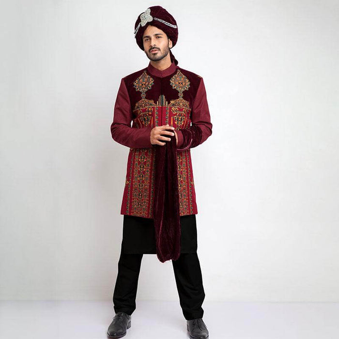 Full-length studio shot of an Arsalan Iqbal deep maroon velvet form-fitted sherwani featuring asymmetrical metallic buttons and a coordinated maroon turban.