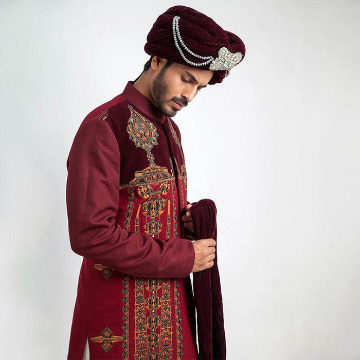 A three-quarter studio portrait of a man in an Arsalan Iqbal cream jamawar sherwani with a gold embroidered mandarin collar, paired with a vibrant red and gold polka dot silk turban and matching shawl.