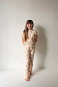 A young girl with long brown hair and glasses stands barefoot against a white wall, smiling gently at the camera in the forever french baby Child Boxy Tee Lounge Set | Lottie—beige, hypo-allergenic kids pajamas with a brown bear print.