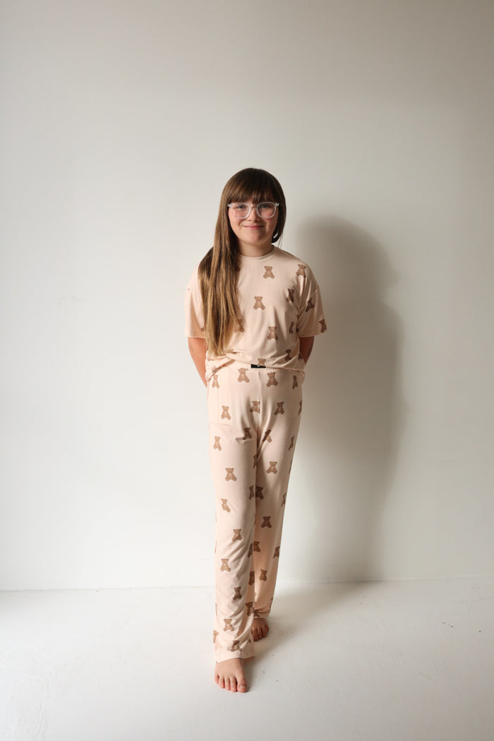 A young girl with long brown hair and glasses stands barefoot against a white wall, smiling gently at the camera in the forever french baby Child Boxy Tee Lounge Set | Lottie—beige, hypo-allergenic kids pajamas with a brown bear print.