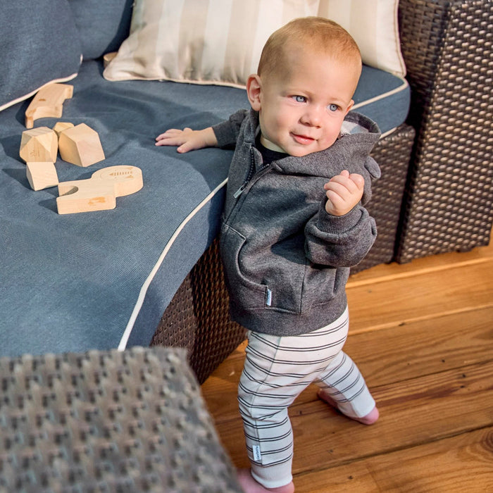 A baby standing on a wooden floor next to a blue sofa, wearing a dark gray zip-up hoodie and white leggings with thin black stripes.