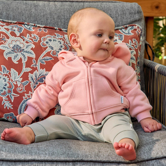A baby sitting on a gray outdoor patio chair wearing a pink zip-up hoodie and light gray pants, with a floral-patterned cushion behind them.