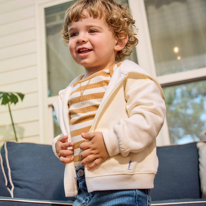 A toddler with curly light brown hair stands on a porch wearing a cream zip-up hoodie over a tan and white striped shirt, paired with blue jeans. The background shows a white house exterior with a blue cushioned sofa.