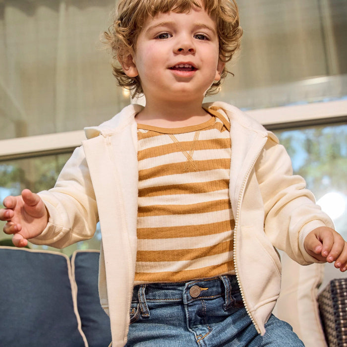 A young child wearing a cream zip-up hoodie over a mustard and cream striped shirt, paired with blue jeans, sitting in a cozy indoor setting.
