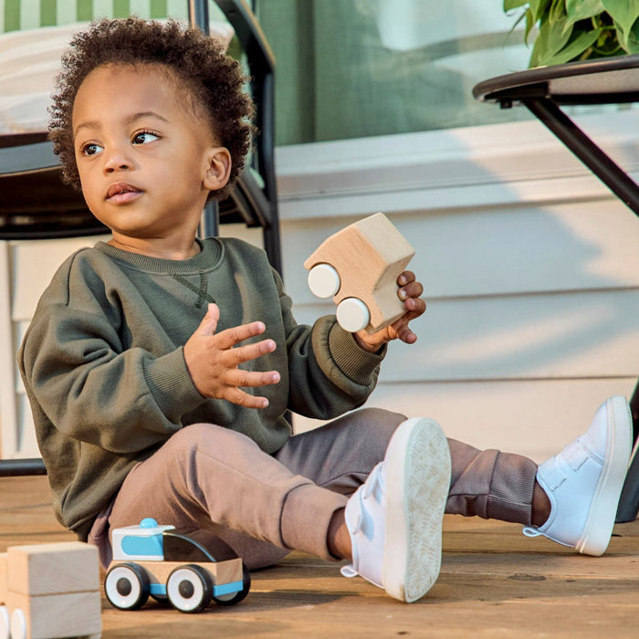 A toddler sits on a wooden deck holding a natural wood toy car, wearing an olive green sweatshirt, brown pants, and white sneakers. Another toy car is in the foreground, with a house exterior and greenery in the background.