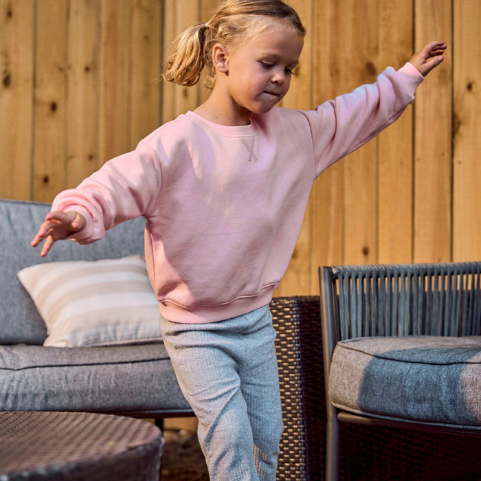A young girl with blonde hair in a ponytail is wearing a light pink crewneck sweatshirt and light gray heathered sweatpants, posing with arms outstretched in front of a wooden wall and patio furniture.