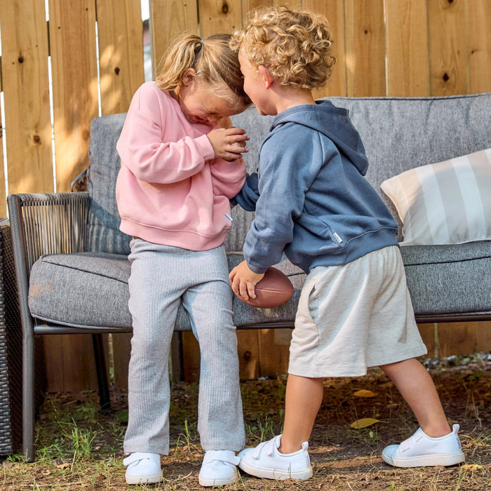 Two children outdoors in front of a gray outdoor sofa. The child on the left wears a pink sweatshirt and light gray sweatpants, and the child on the right wears a slate blue hoodie, light beige shorts, and white sneakers.