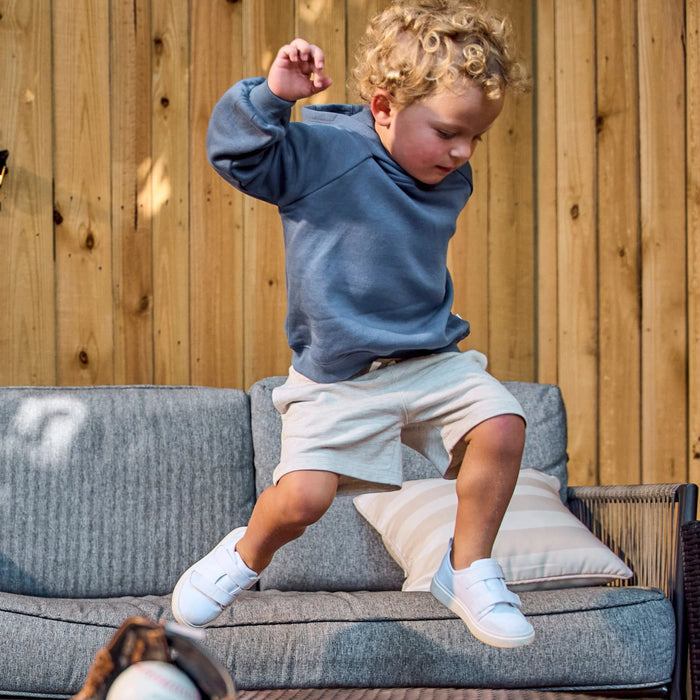 A young child with curly blonde hair wearing a slate blue hoodie, cream shorts, and white sneakers jumps onto a gray outdoor sofa in front of a wooden fence.