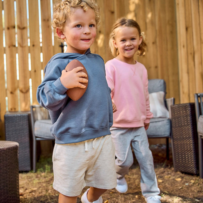A young boy in a slate blue hoodie and beige shorts holds a football while smiling, with a young girl in a pink sweatshirt and gray sweatpants walking in the background in front of a wooden fence.