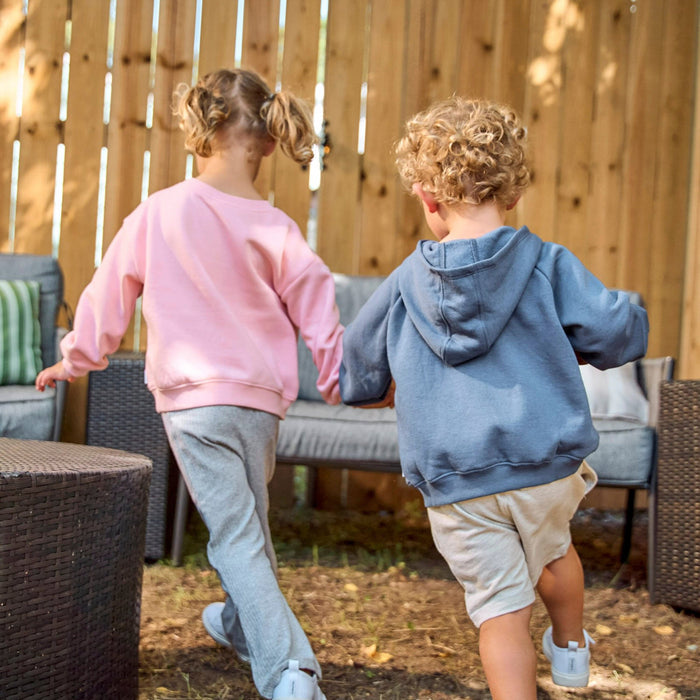Two children walking away in an outdoor backyard setting. The girl wears a pink sweatshirt and light gray sweatpants, and the boy wears a dusty blue hoodie, cream shorts, and white sneakers.