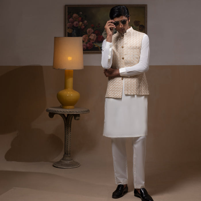 Arsalan Iqbal Light Gold Embroidered Waistcoat with IronEZ Kurta & Pyjama Set man wearing a light gold heavily embroidered sleeveless waistcoat with gold button closure over a crisp white full-sleeve kurta, paired with matching white straight-cut pyjama, accessorized with sunglasses, styled in a warm classic indoor setting