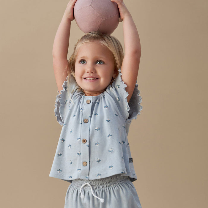 Child wearing a light blue top with ruffled sleeves and patterned pants, holding a pink ball above their head against a beige background.