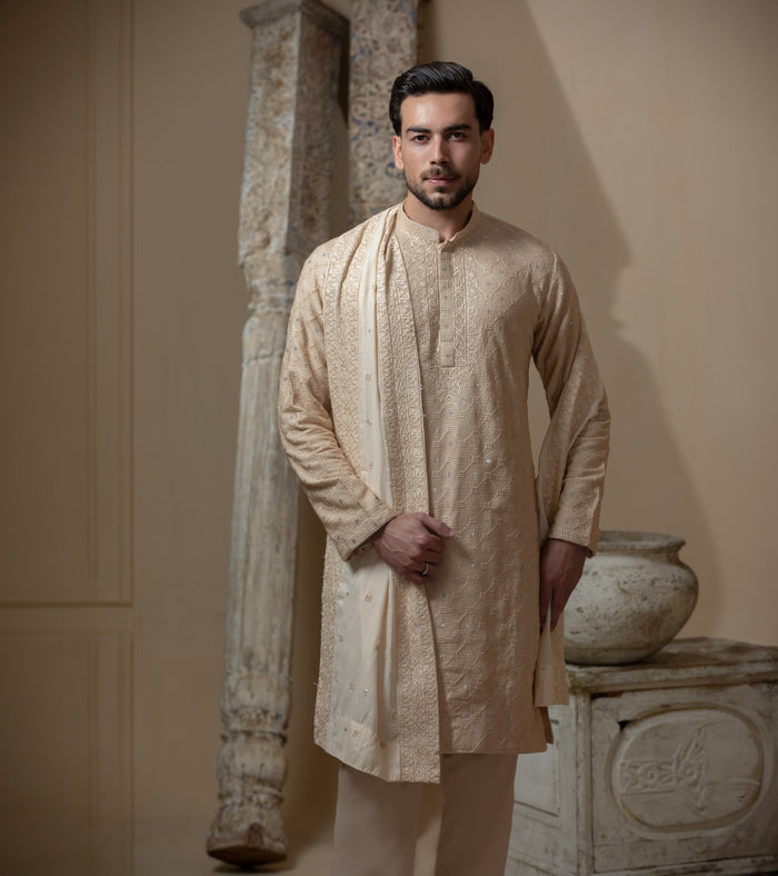 Man wearing Arsalan Iqbal's Peach & Gold Chikankari Kurta Pyjama with a cream stole draped over shoulders, styled with dark formal shoes against a warm beige backdrop