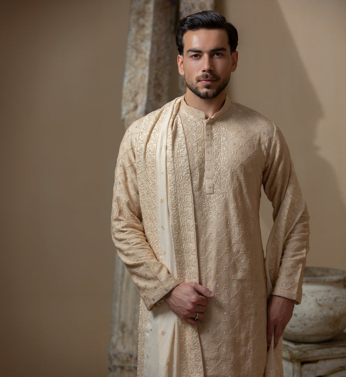 Man wearing Arsalan Iqbal's Peach & Gold Chikankari Kurta Pyjama with a cream stole draped over shoulders, styled with dark formal shoes against a warm beige backdrop