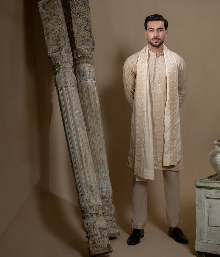 Man wearing Arsalan Iqbal's Peach & Gold Chikankari Kurta Pyjama with a cream stole draped over shoulders, styled with dark formal shoes against a warm beige backdrop