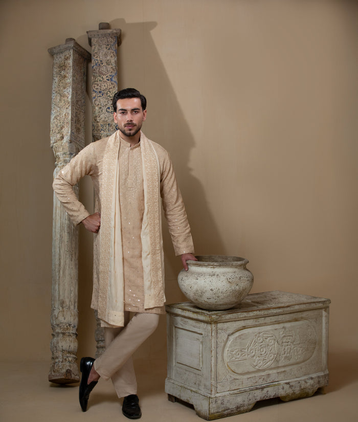 Man wearing Arsalan Iqbal's Peach & Gold Chikankari Kurta Pyjama with a cream stole draped over shoulders, styled with dark formal shoes against a warm beige backdrop