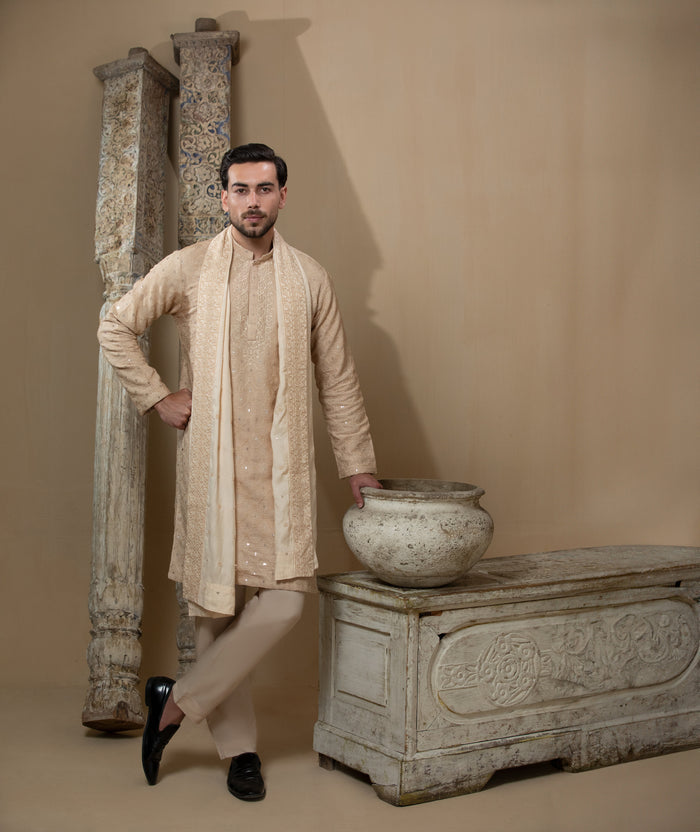 Man wearing Arsalan Iqbal's Peach & Gold Chikankari Kurta Pyjama with a cream stole draped over shoulders, styled with dark formal shoes against a warm beige backdrop