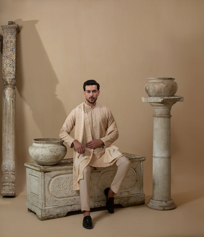 Man wearing Arsalan Iqbal's Peach & Gold Chikankari Kurta Pyjama with a cream stole draped over shoulders, styled with dark formal shoes against a warm beige backdrop