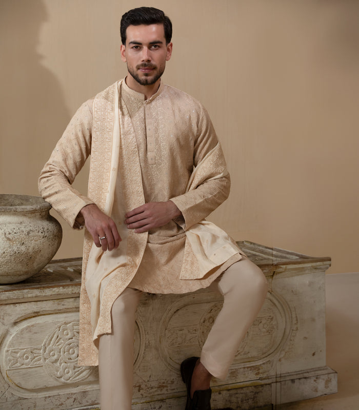 Man wearing Arsalan Iqbal's Peach & Gold Chikankari Kurta Pyjama with a cream stole draped over shoulders, styled with dark formal shoes against a warm beige backdrop