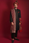 Full-length portrait of Arsalan Iqbal luxury black sherwani featuring gold buttons, paired with a matching maroon silk turban and an intricately embroidered maroon silk shawl with gold motifs, styled with black trousers and loafers.