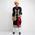 Full-length studio shot of a man in an Arsalan Iqbal black velvet sherwani featuring large, ornate gold paisley embroidery on the chest and hem, paired with a gold turban and a draped red shawl.