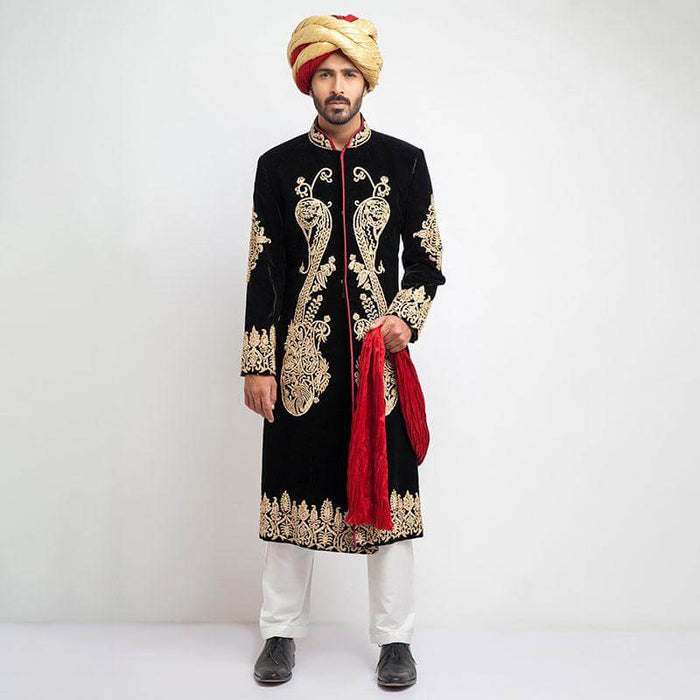 Full-length studio shot of a man in an Arsalan Iqbal black velvet sherwani featuring large, ornate gold paisley embroidery on the chest and hem, paired with a gold turban and a draped red shawl.