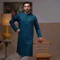 Arsalan Iqbal Teal Blue Chikankari Kurta Pyjama  man wearing a rich teal blue full-sleeve kurta with subtle all-over chikankari embroidery, mandarin collar with button placket, paired with white straight-cut pyjama, posed confidently with one hand on a wooden chair, styled in a warm classic indoor setting with a yellow vase and floral painting in the background