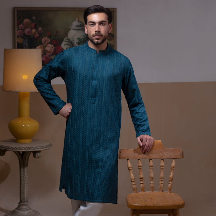 Arsalan Iqbal Teal Blue Chikankari Kurta Pyjama  man wearing a rich teal blue full-sleeve kurta with subtle all-over chikankari embroidery, mandarin collar with button placket, paired with white straight-cut pyjama, posed confidently with one hand on a wooden chair, styled in a warm classic indoor setting with a yellow vase and floral painting in the background