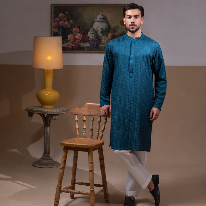 Arsalan Iqbal Teal Blue Chikankari Kurta Pyjama  man wearing a rich teal blue full-sleeve kurta with subtle all-over chikankari embroidery, mandarin collar with button placket, paired with white straight-cut pyjama, posed confidently with one hand on a wooden chair, styled in a warm classic indoor setting with a yellow vase and floral painting in the background