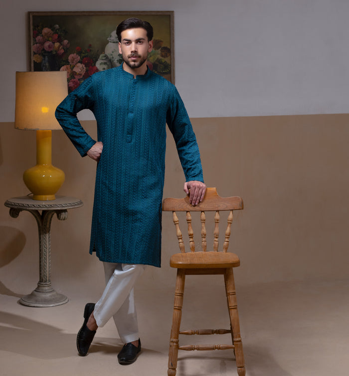 Arsalan Iqbal Teal Blue Chikankari Kurta Pyjama  man wearing a rich teal blue full-sleeve kurta with subtle all-over chikankari embroidery, mandarin collar with button placket, paired with white straight-cut pyjama, posed confidently with one hand on a wooden chair, styled in a warm classic indoor setting with a yellow vase and floral painting in the background
