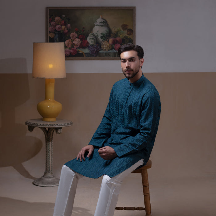 Arsalan Iqbal Teal Blue Chikankari Kurta Pyjama  man wearing a rich teal blue full-sleeve kurta with subtle all-over chikankari embroidery, mandarin collar with button placket, paired with white straight-cut pyjama, posed confidently with one hand on a wooden chair, styled in a warm classic indoor setting with a yellow vase and floral painting in the background