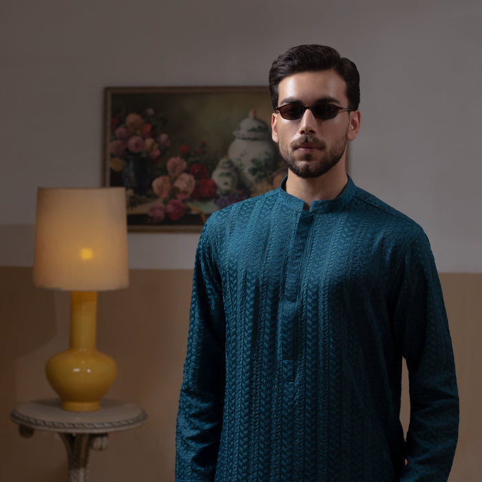 Arsalan Iqbal Teal Blue Chikankari Kurta Pyjama  man wearing a rich teal blue full-sleeve kurta with subtle all-over chikankari embroidery, mandarin collar with button placket, paired with white straight-cut pyjama, posed confidently with one hand on a wooden chair, styled in a warm classic indoor setting with a yellow vase and floral painting in the background