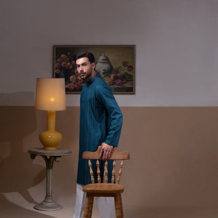 Arsalan Iqbal Teal Blue Chikankari Kurta Pyjama man wearing a rich teal blue full-sleeve kurta with subtle all-over chikankari embroidery, mandarin collar with button placket, paired with white straight-cut pyjama, posed confidently with one hand on a wooden chair, styled in a warm classic indoor setting with a yellow vase and floral painting in the background