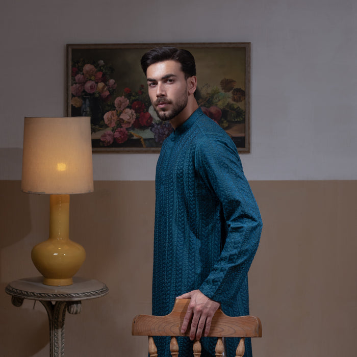 Arsalan Iqbal Teal Blue Chikankari Kurta Pyjama – man wearing a rich teal blue full-sleeve kurta with subtle all-over chikankari embroidery, mandarin collar with button placket, paired with white straight-cut pyjama, posed confidently with one hand on a wooden chair, styled in a warm classic indoor setting with a yellow vase and floral painting in the background