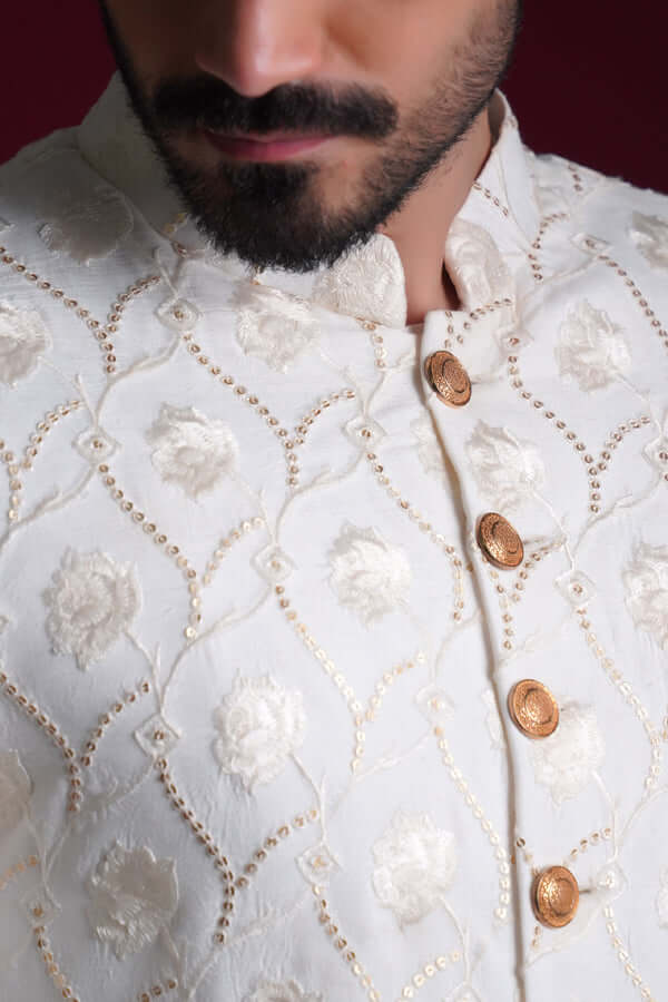 Extreme close-up of Arsalan Iqbal ivory silk sherwani featuring raised white floral embroidery, gold-toned textured buttons, and intricate sequin-lined vine patterns.